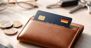 Nahaufnahme einer Kreditkarte mit deutscher Flagge, die in einer braunen Leder-Geldbörse steckt. Daneben liegen Euro-Münzen auf einem Holztisch, im Hintergrund sind unscharf ein Autoschlüssel und eine Brille zu sehen. Das Bild steht für bargeldloses Bezahlen, Kreditkartennutzung in Deutschland und persönliche Finanzorganisation im Alltag.